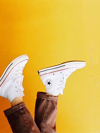 Person wearing brown pants and white sneakers with legs raised upward against a bright yellow background, creating a playful, energetic and minimalist lifestyle scene.