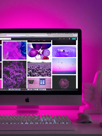 Desktop computer displaying image gallery on screen with pink and purple neon lighting, keyboard and decor visible, creating a modern creative workspace aesthetic.